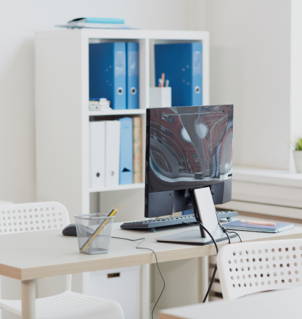 A clean office desk with a computer monitor, keyboard, mouse, and stationery. Behind the desk is a white shelf with blue and white binders. The scene suggests a neat and organized work environment.